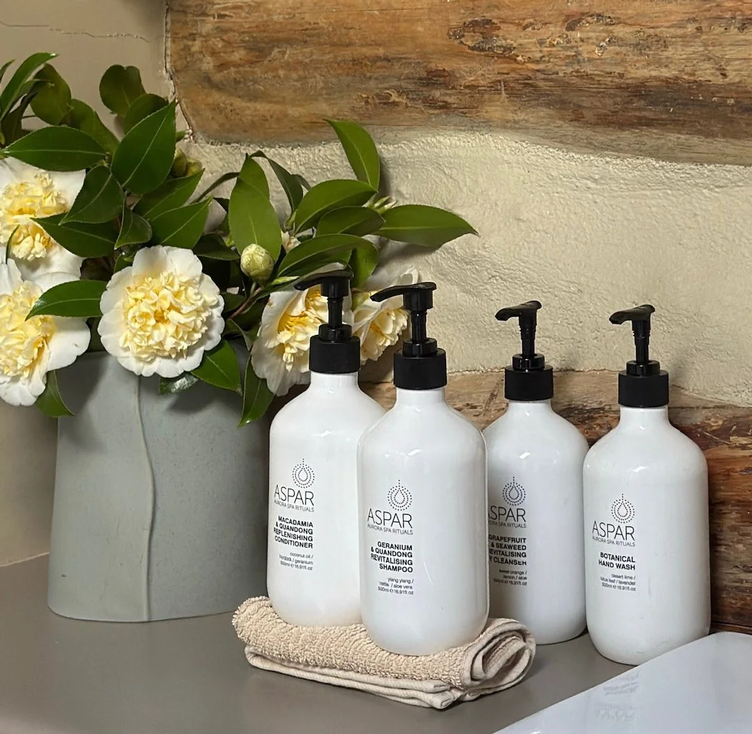 A set of four white bottles with black pump dispensers labeled as skincare products, placed on a folded beige towel next to a gray vase filled with white and yellow flowers, against a rustic wooden background.