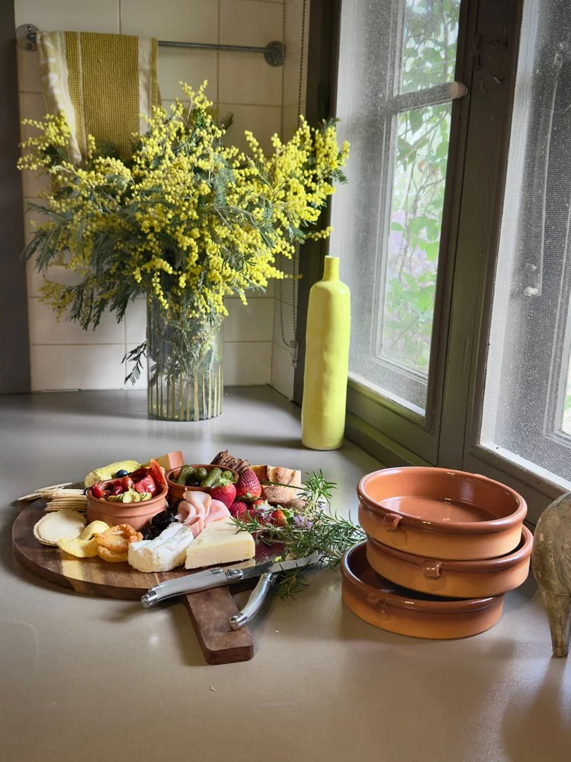 Kitchen windowsill with a vase of yellow flowers, a yellow vase, terracotta dishes, a cheese platter with berries, grapes, and slices of meat and cheese, a cutting board, cheese knives, and a sprig of herbs.