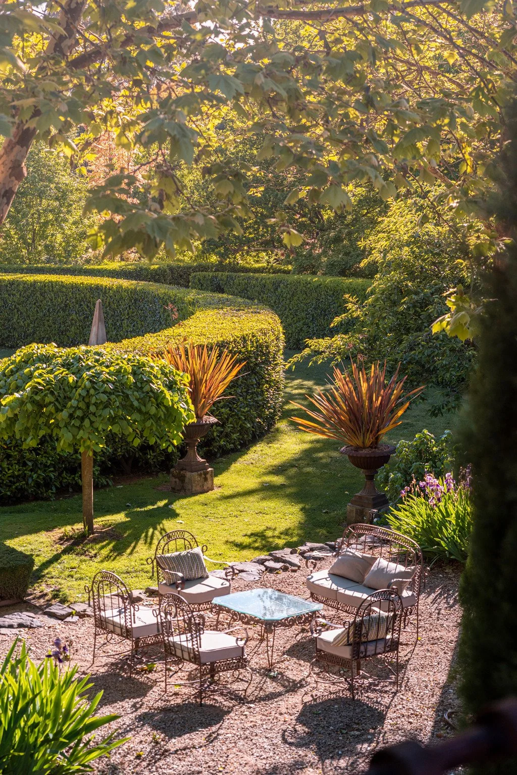 A cozy outdoor garden seating area with a glass table and wrought iron chairs surrounded by lush green bushes, plants, and trees on a sunny day.