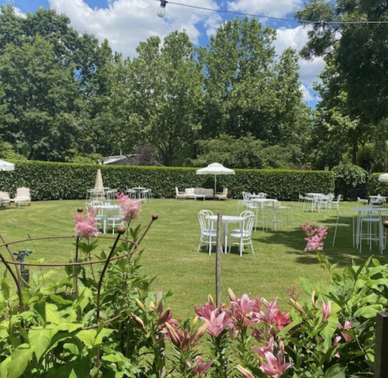 Outdoor garden with green grass, white patio furniture, pink flowers in the foreground, and tall trees under a partly cloudy sky.