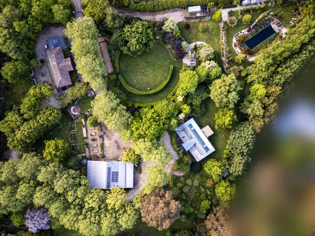 Aerial view of Planetrees Estate, a lush green yard with multiple structures, including a house with solar panels, a swimming pool, and a large circular lawn area surrounded by trees.