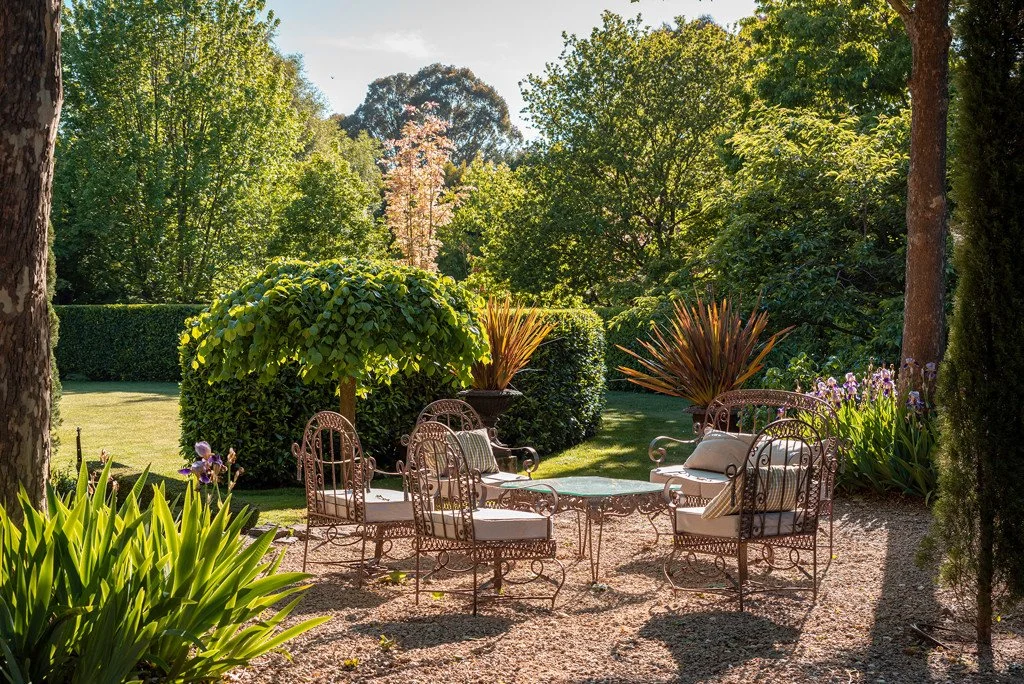 A garden patio with wrought iron furniture including chairs and a round table, surrounded by lush green trees, bushes, and colorful plants, under a clear blue sky.