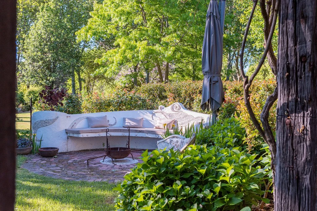 Outdoor garden seating area with a curved white concrete bench, pastel cushions, a black umbrella, and a fire pit, surrounded by lush greenery including shrubs, trees, and flowering plants.