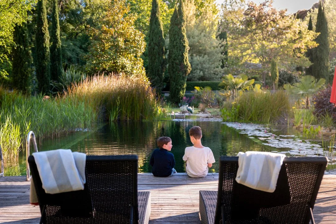 Two children sitting on a dock by Planetrees Estate natural pool, surrounded by lush greenery and tall trees, with chairs and towels in the foreground.