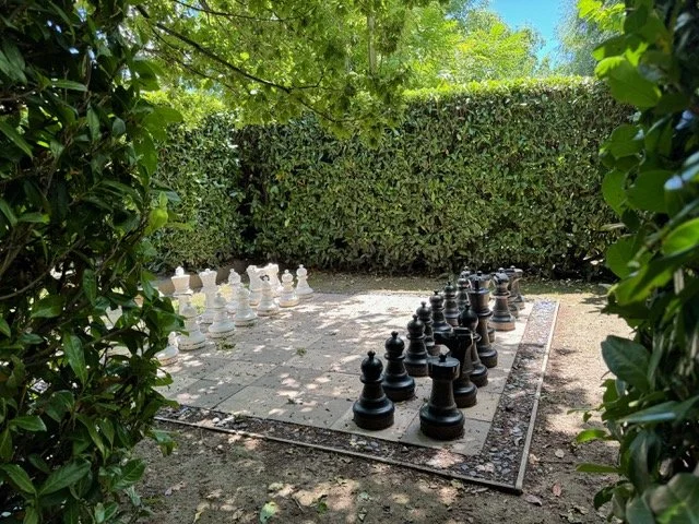 An outdoor giant chessboard with large black and white chess pieces, surrounded by green bushes and trees on a sunny day.