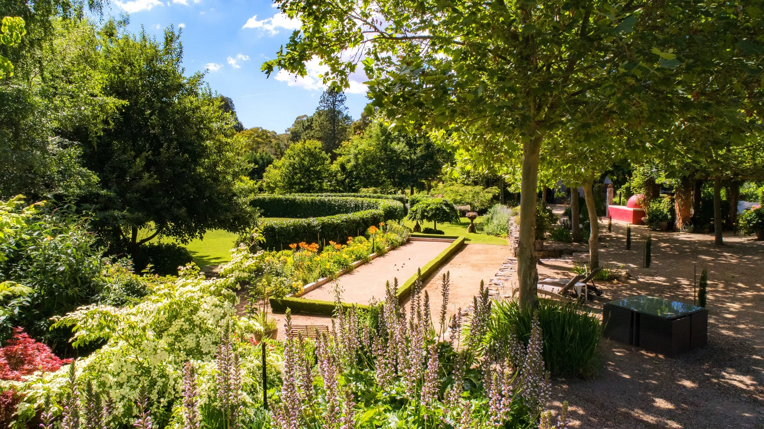 A lush garden at Planetrees Estate with various green trees and bushes, colourful flowers, a sandy pathway, and benches under shaded areas on a sunny day.