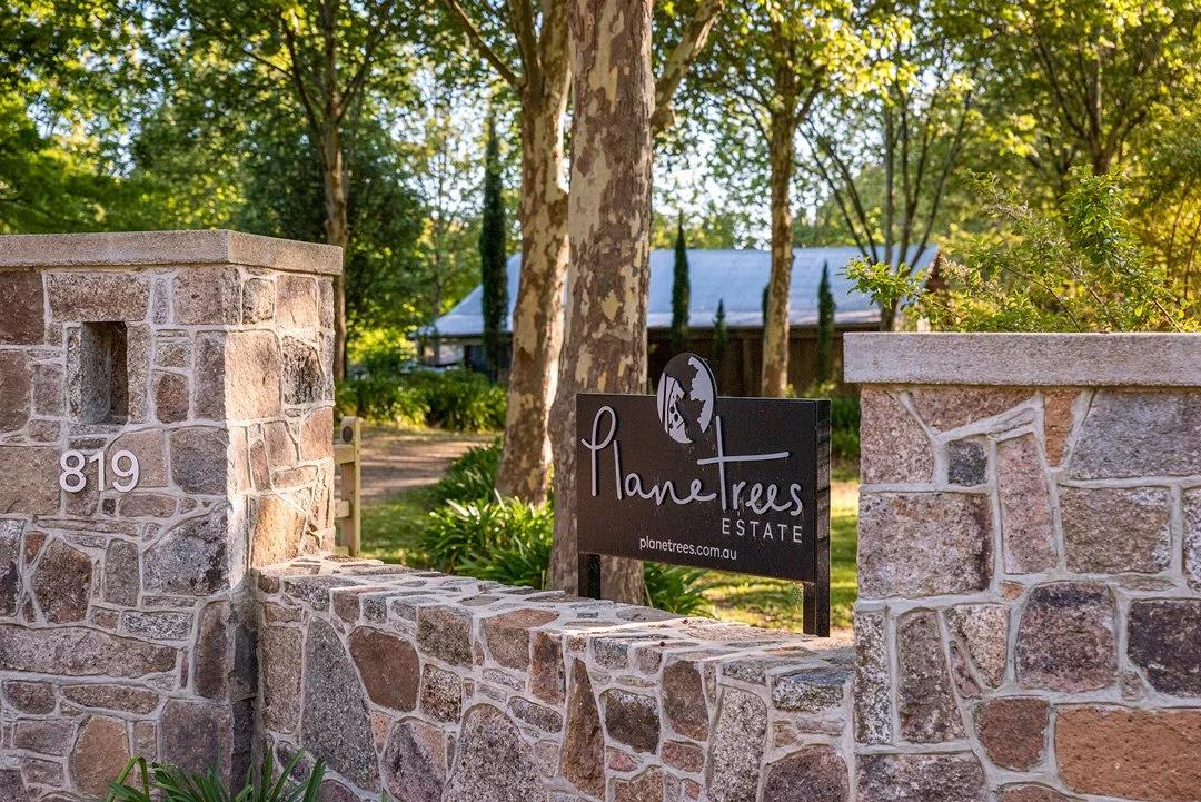 Stone entrance with a sign reading "Planetrees Estate" and the website "planetrees.com.au." A tree, a house, and greenery are visible in the background.