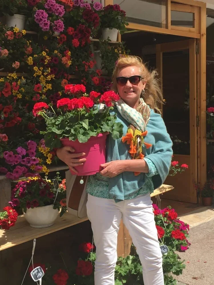 Planetrees Estate owner, Genevieve Milham, holding a pink pot of red flowers in front of a flower shop with various colourful flowers on display.