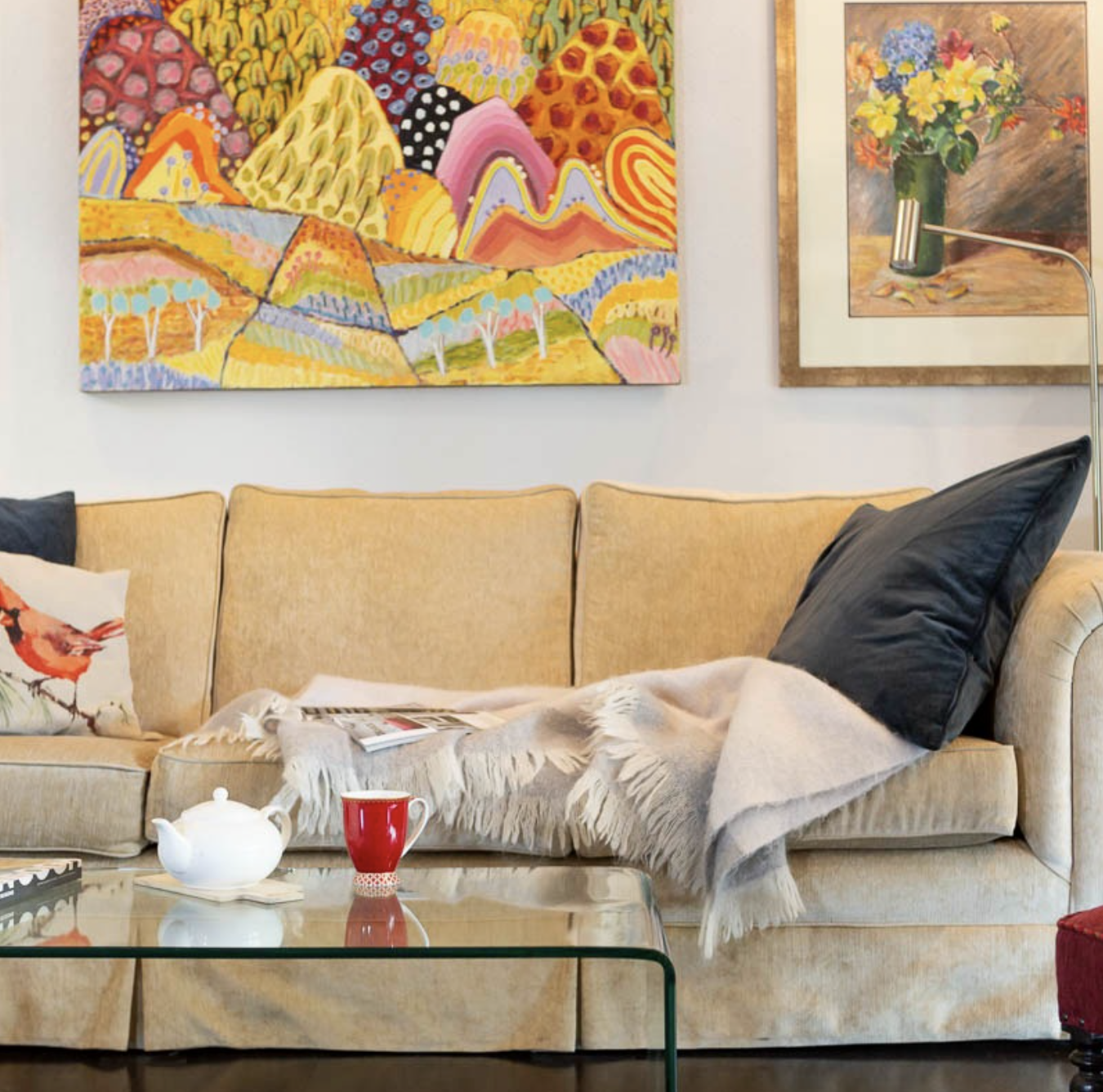 Living room with a beige sofa, black pillow, floral and bird throw pillows, a glass coffee table with a white teapot and red mug, colorful abstract and floral art on the wall, and a floor lamp.