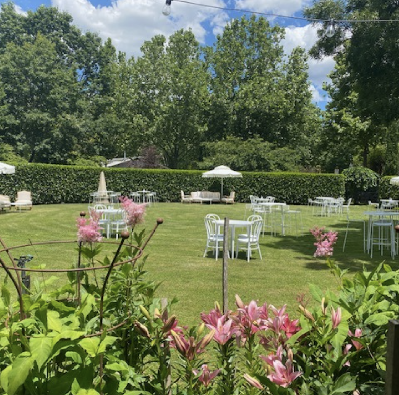 An outdoor garden with white tables and chairs, pink flowers, and green trees under a partly cloudy sky.