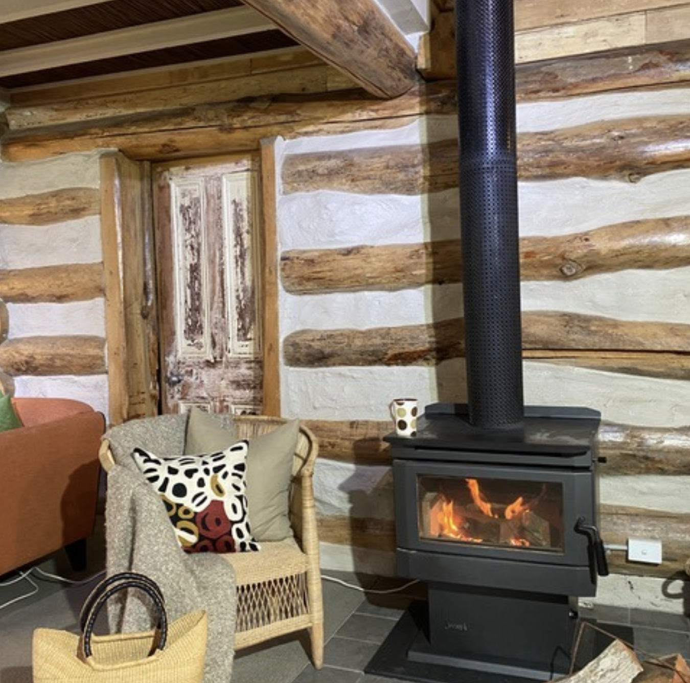 Living room corner with a wood stove, fire burning inside, a wicker chair with a colorful patterned pillow, a beige pillow, a yellow woven basket, and a wall made of horizontal wooden logs with white plaster between them.