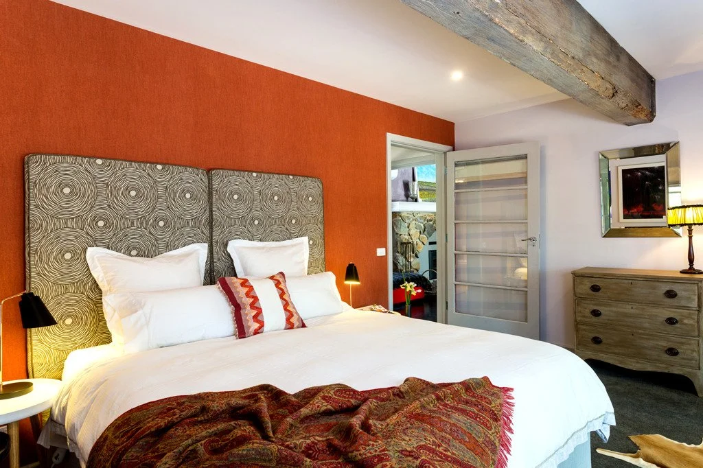 A cozy bedroom with a large bed, patterned headboard, and red accent wall. There are three pillows and a throw blanket on the bed. A doorway opens to an outside area with rocks. A dresser with a mirror, a lamp, and wall decor are visible.