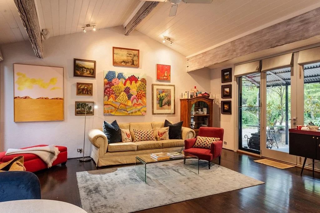 A cozy living room with a beige sofa, a red armchair, and a black cushion. Artworks adorn a white wall behind the sofa, and a glass coffee table is in front. There is a large sliding glass door leading outside, revealing greenery, allowing natural li