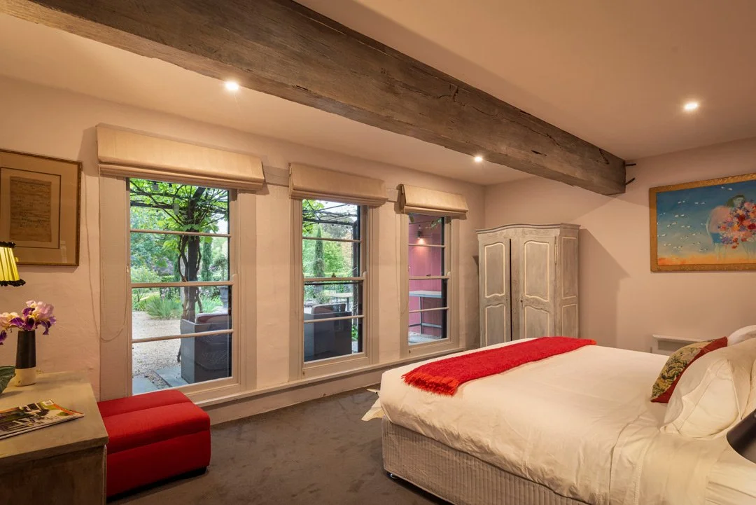Bedroom with large windows, a bed with white linens and a red throw, a wooden armoire, and outdoor seating visible through the windows.
