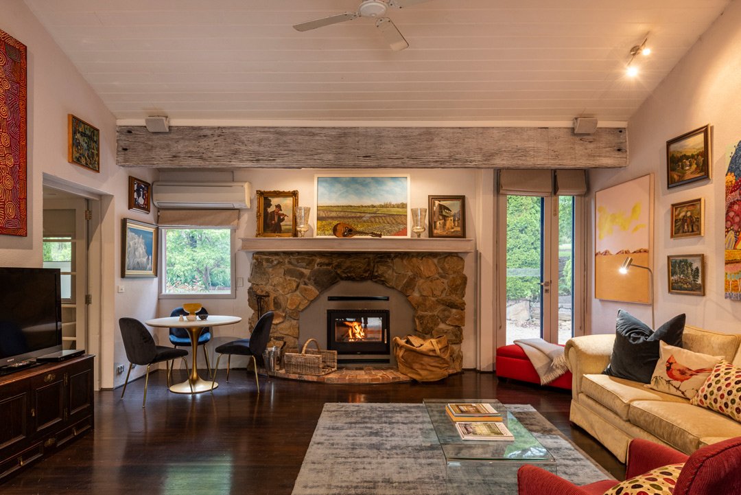Living room with a stone fireplace, sofas, a glass coffee table, artwork on the walls, and a large window and glass door showing greenery outside.