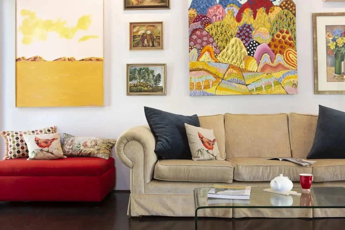 Living room with a beige sofa, a red chaise, and various pillows, including one with a robin. Wall decorated with colorful paintings and framed art, a glass coffee table with books, a teapot, and a cup.