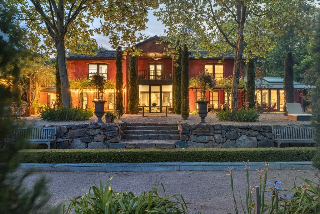 A large house at Planetrees Estate illuminated at dusk, with a garden and stone steps leading to a patio area, surrounded by trees and greenery.