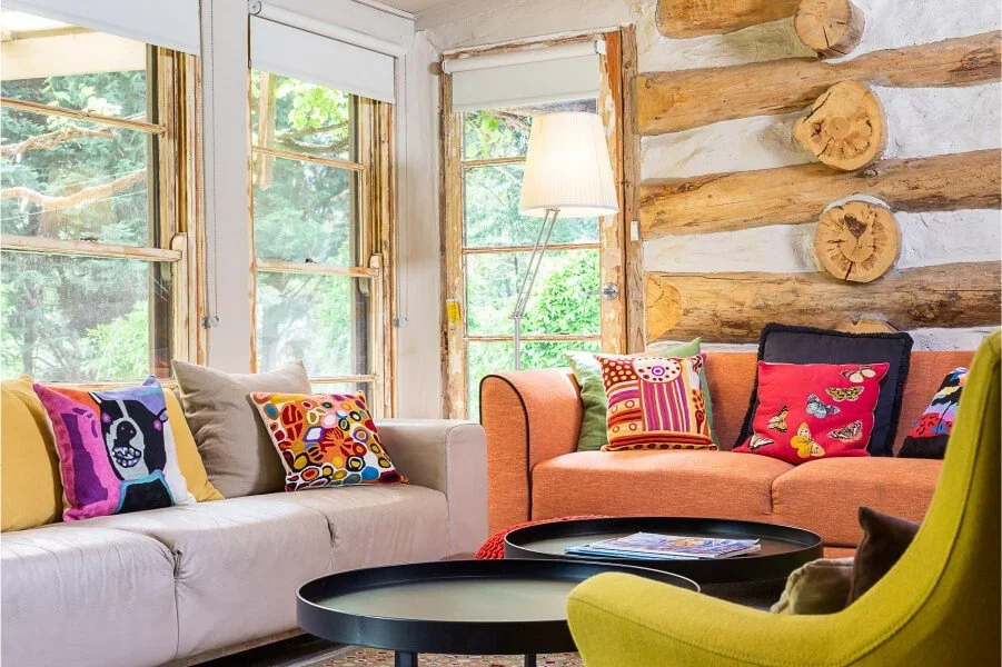 Living room with three sofas: one beige, one orange, and one green, decorated with colorful pillows featuring various patterns and characters. The room has large windows with views of green trees outside. A standing lamp with a white shade is near the window. The wall behind the sofas is made of logs with visible wood ends.