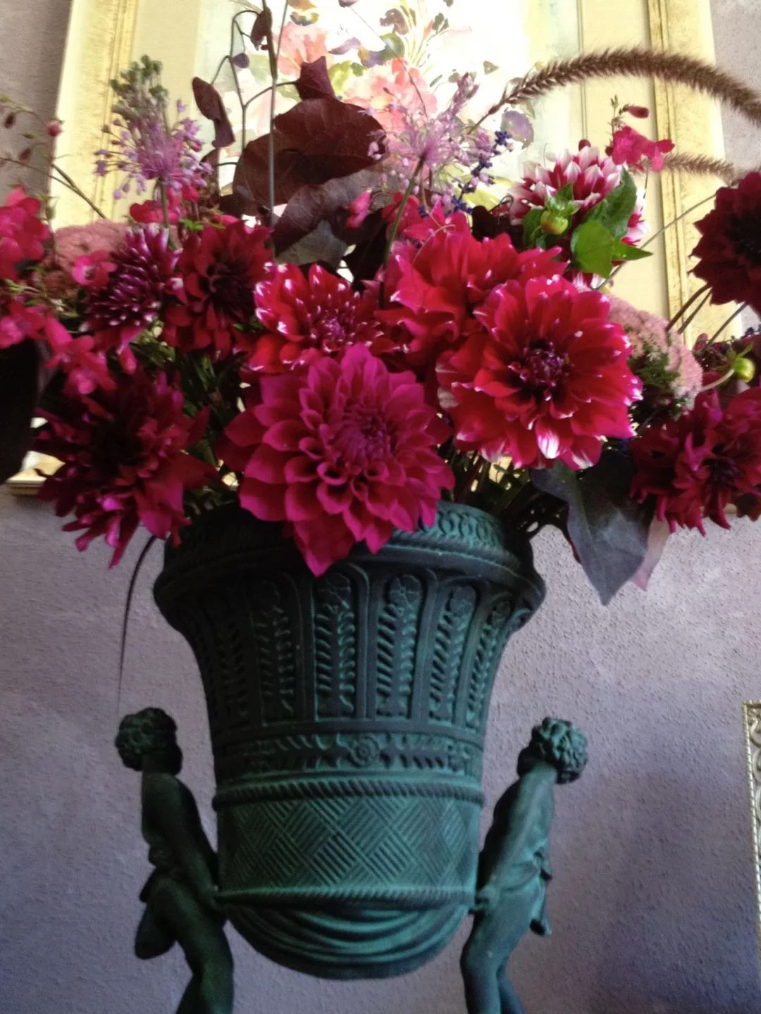 Arrangement of bright pink and red flowers in a decorative dark green vase with two small statues of figures on either side, set against a purple wall and a framed picture in the background.