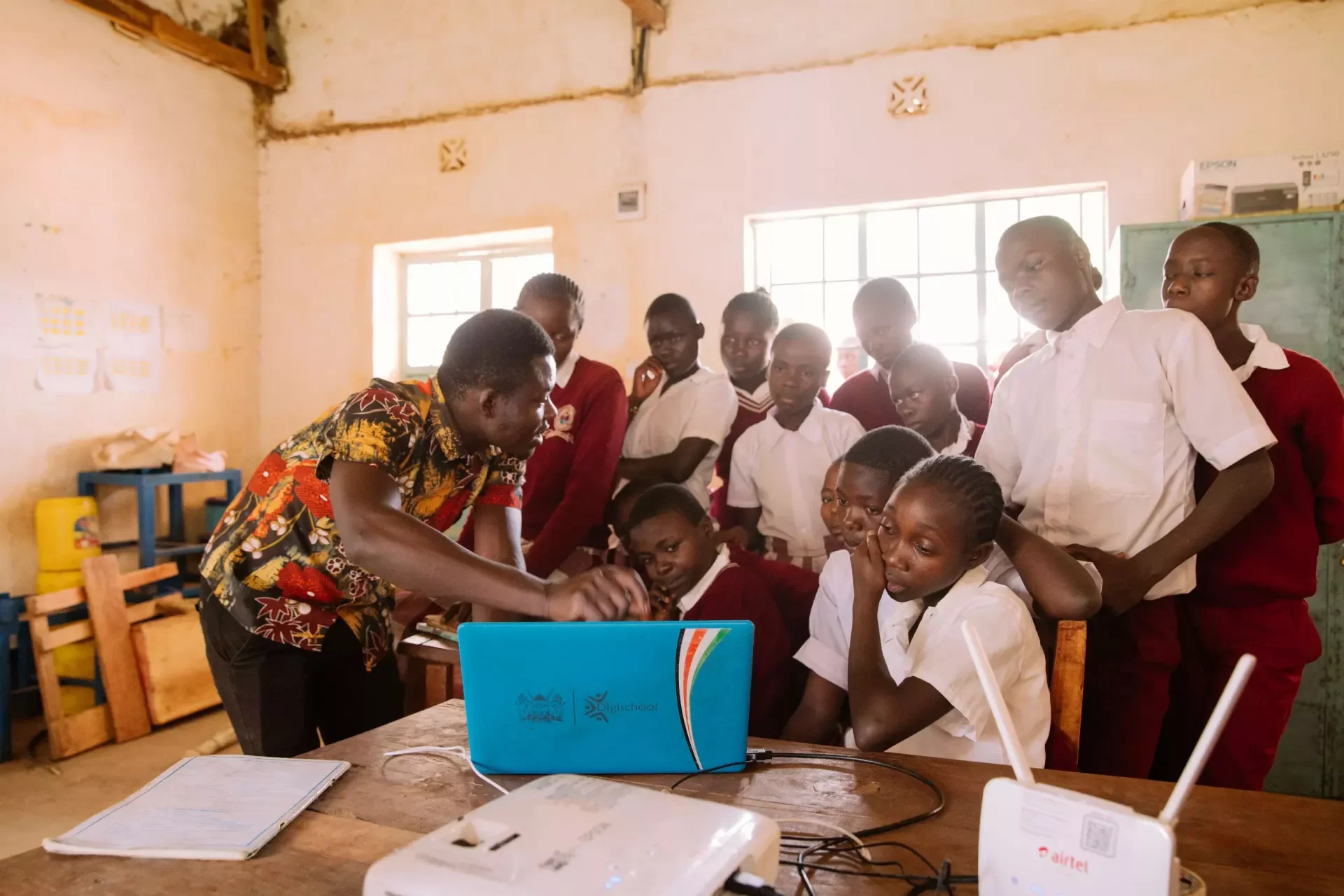 An instructor demonstrates something on a laptop to a group of students in a classroom with worn walls, a window, and some classroom supplies.