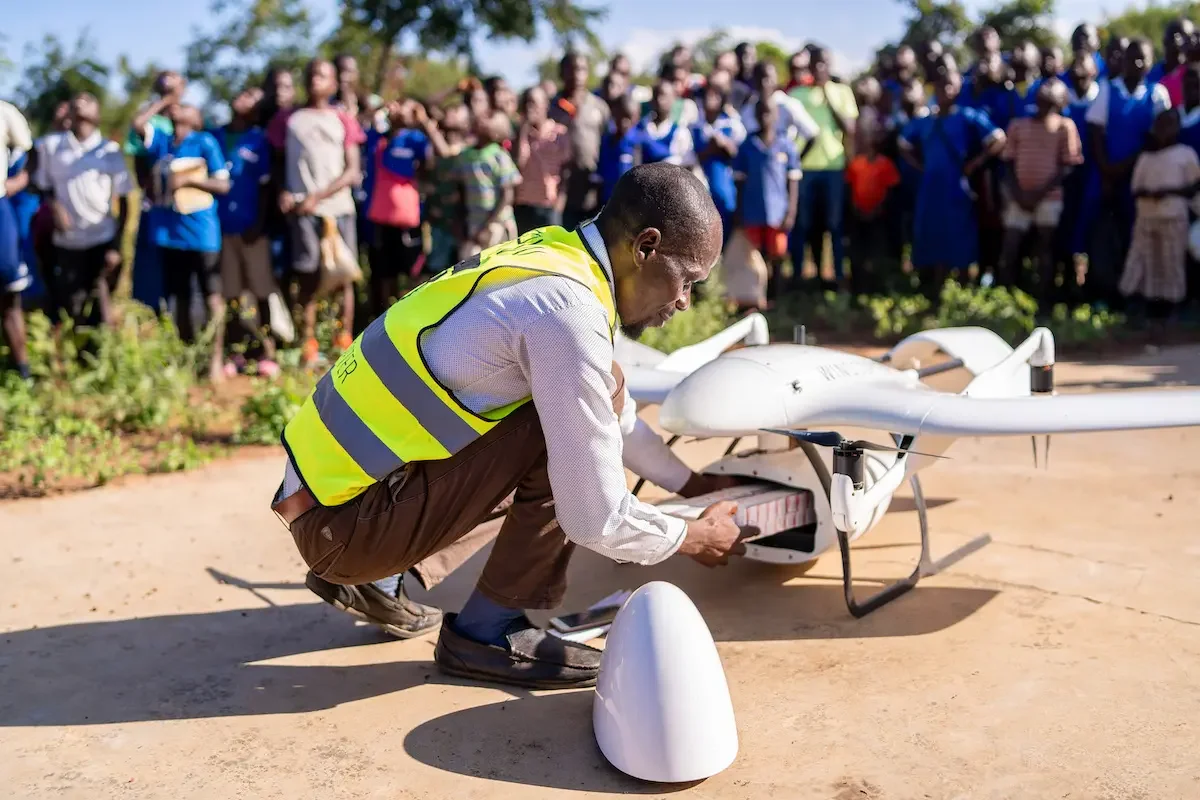 A man in a yellow safety vest kneeling as he prepares a white drone for flight in front of a large group of people outdoors on a sunny day.