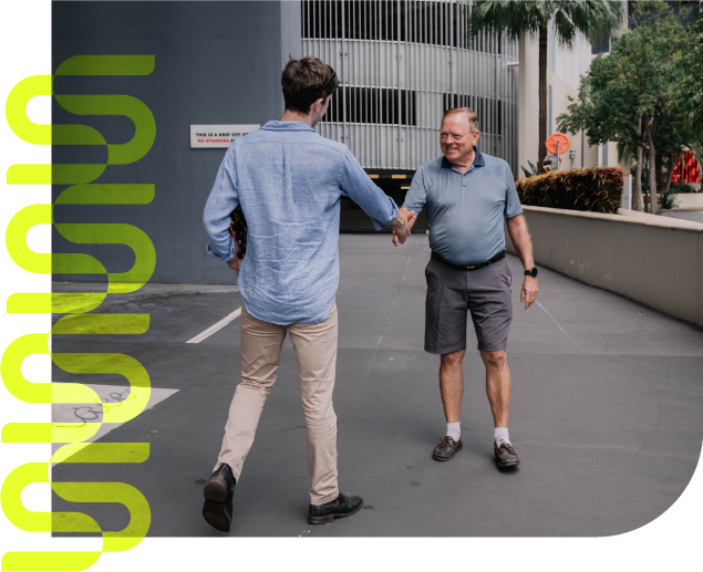 Two men shake hands in a parking lot near a modern building, smiling as they greet each other.