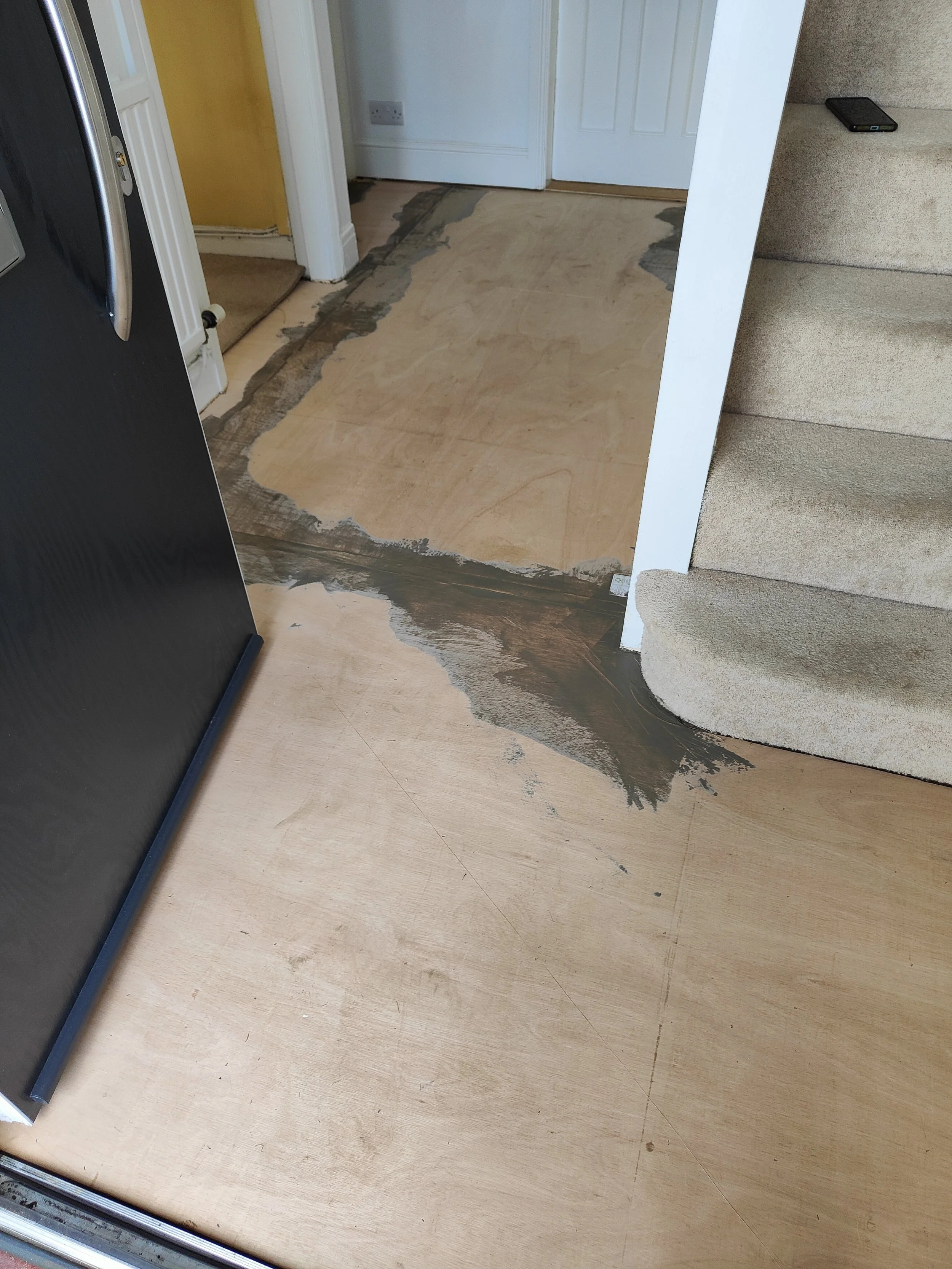 Part of a home interior showing a staircase on the right, a black refrigerator on the left, a yellow painted wall in the background, and flooring that is undergoing renovation or repair, with parts of the original floor removed and exposed.