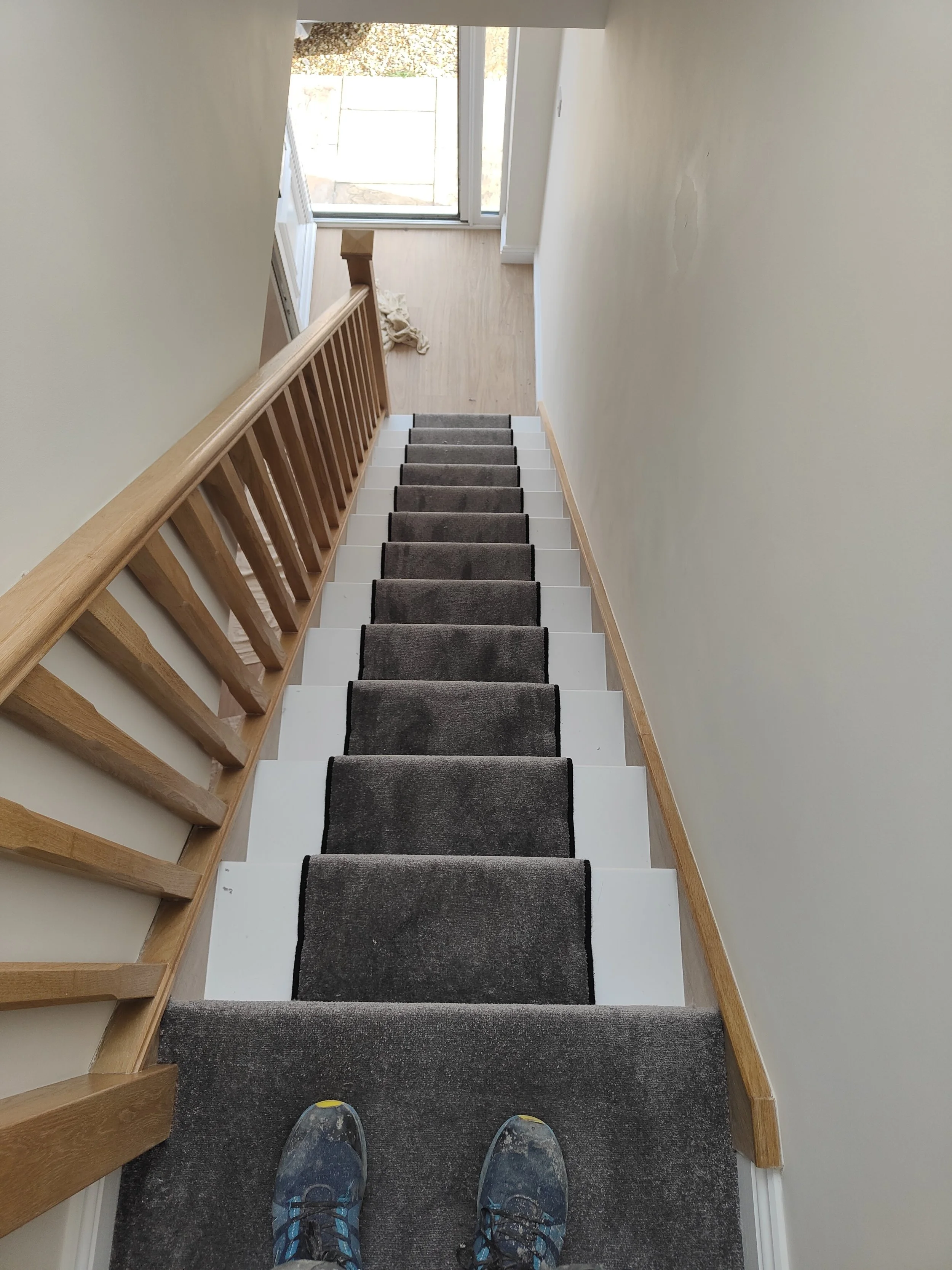 A view looking down a staircase with a dark gray carpet runner and wooden handrails, ending at a small landing with a sliding glass door that leads outside.