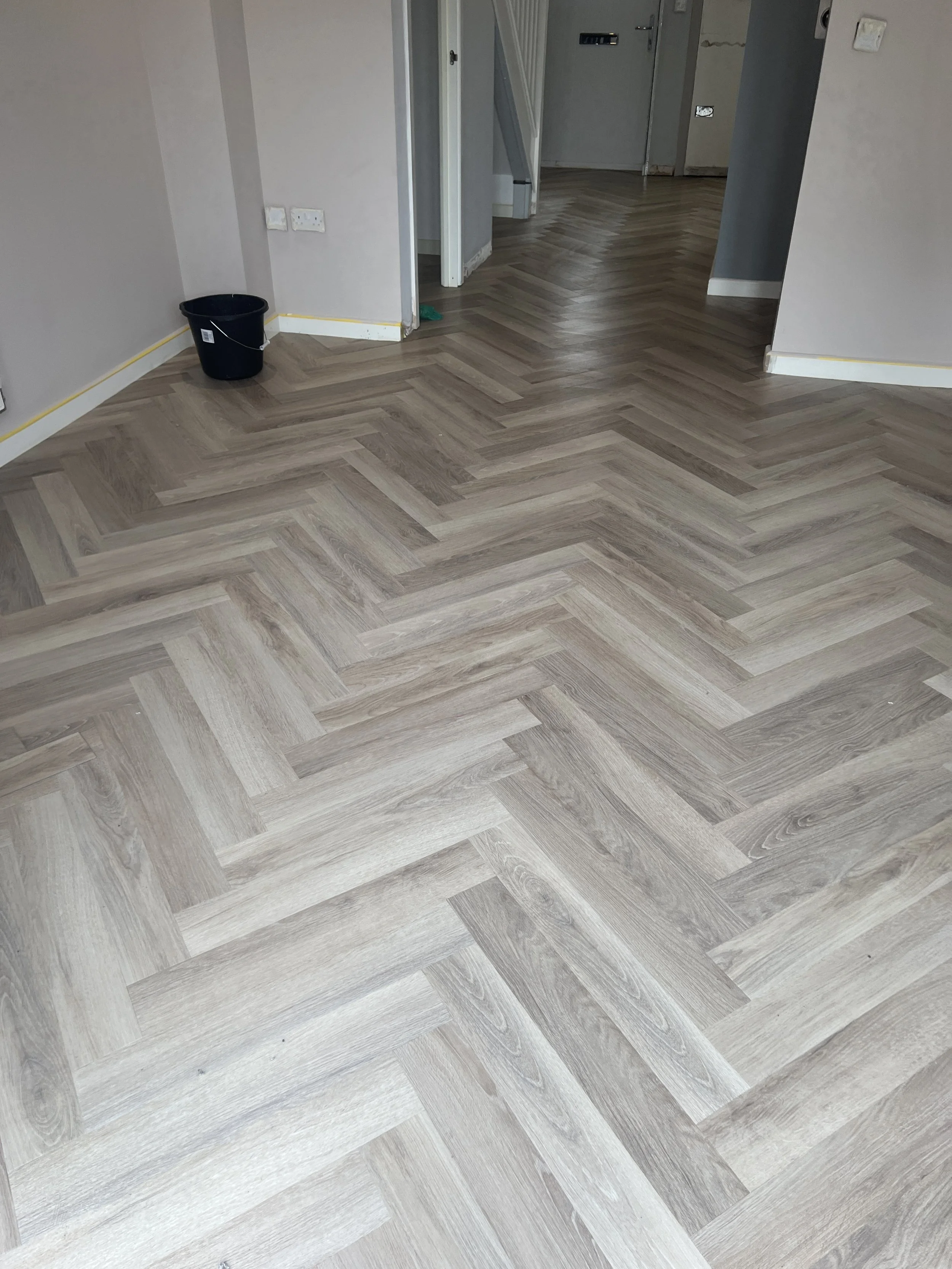Room with newly installed light wood herringbone pattern flooring, a small black bucket near wall, and unfinished walls with visible electrical outlets and switches.