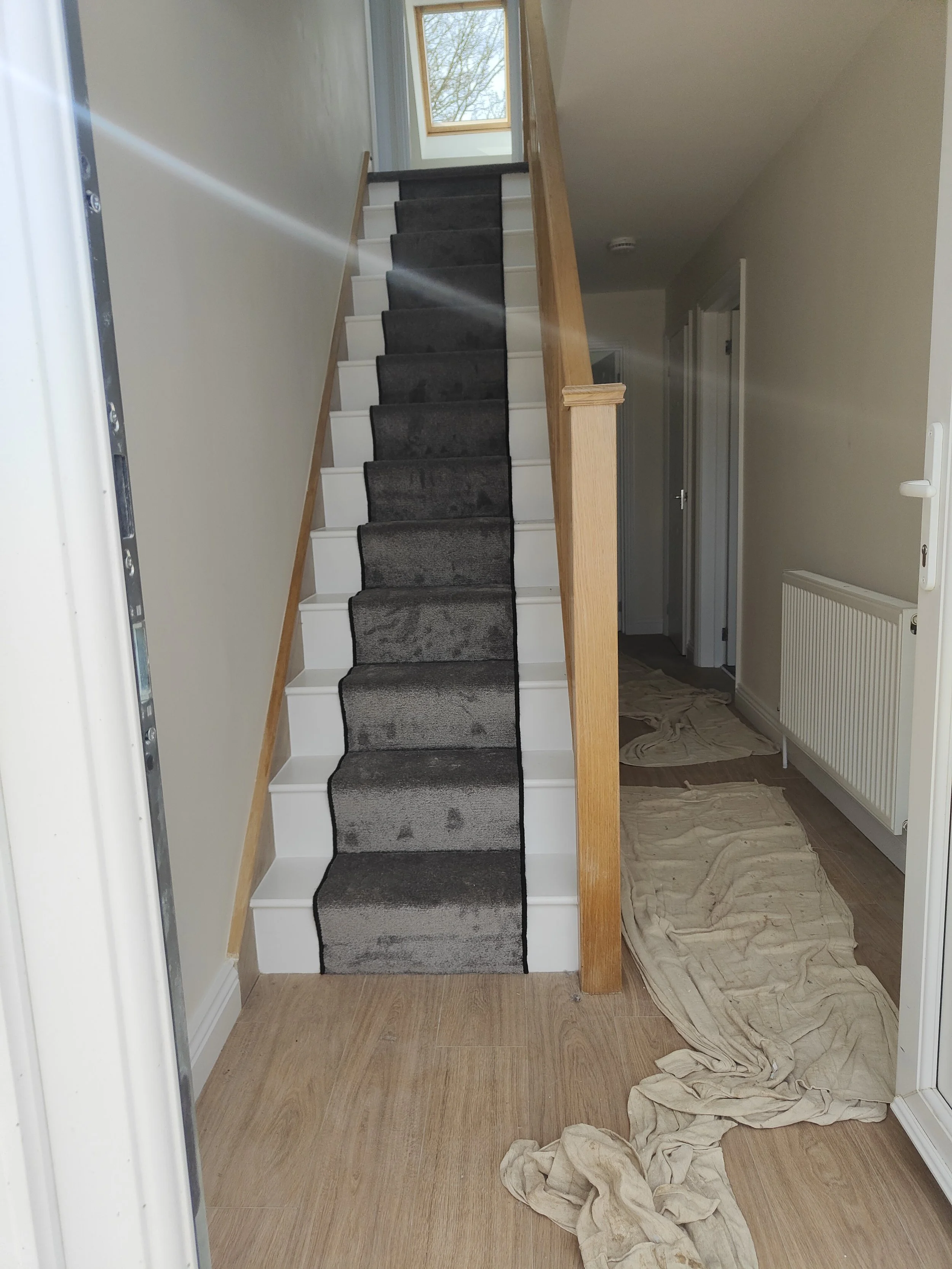 Interior view of a stairway under renovation, with a new black carpet runner on the stairs, scattered drop cloths on the wooden floor, and an open hallway with a radiator and doorway.