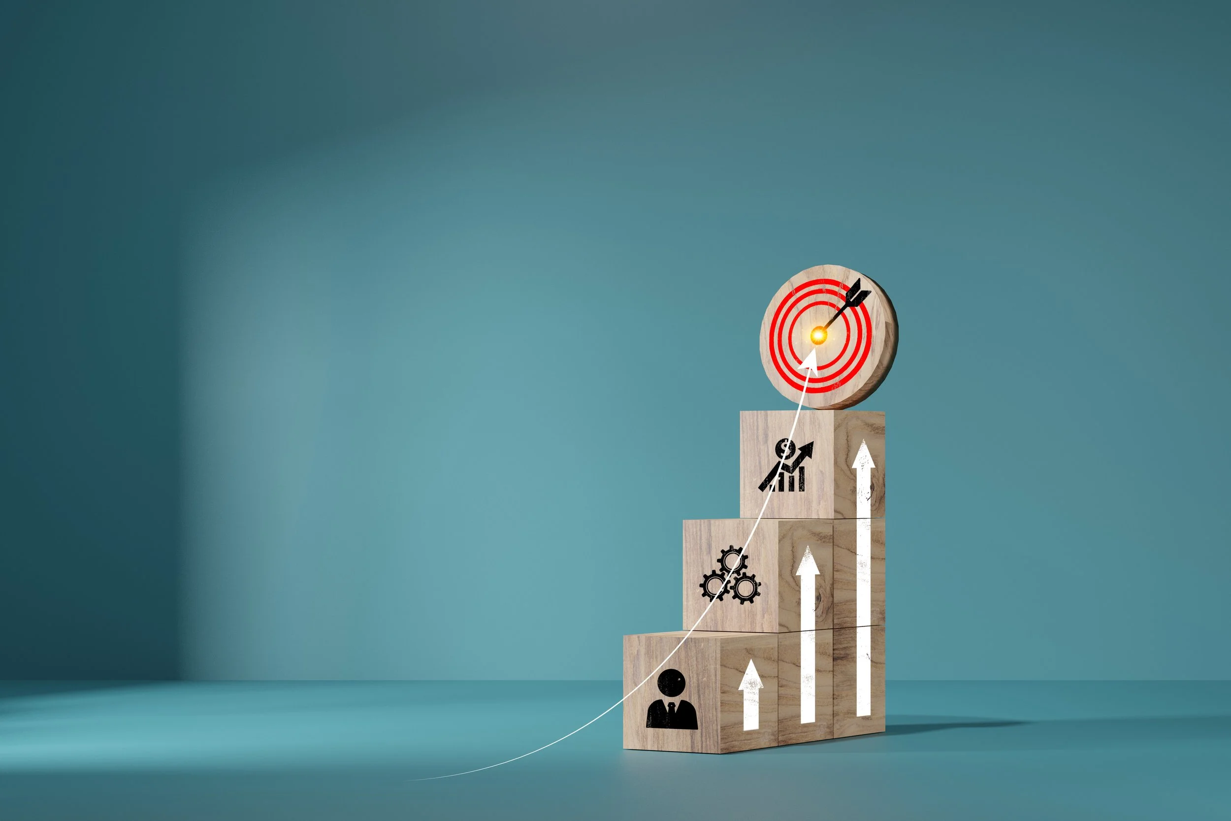 Wooden blocks stacked in a stair-step pattern with icons and arrows, topped by a target with an arrow hitting the bullseye. The blocks feature icons of a person, gears, and a chart with an upward arrow, representing growth and achievement.