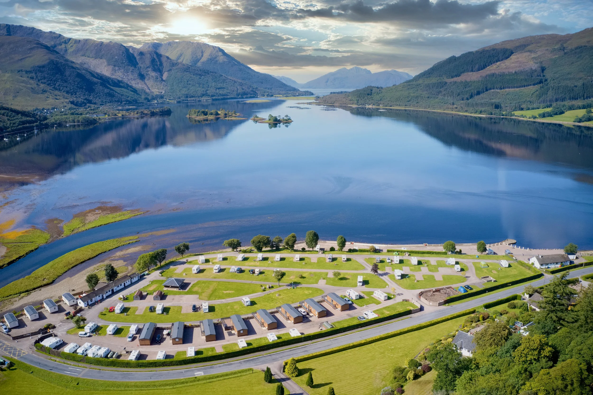 Aerial view of a large lake surrounded by mountains. In the foreground, there is a caravan park with numerous RVs and small buildings, with a well-maintained grassy area and trees. The water is calm and reflects the sky and terrain around it.