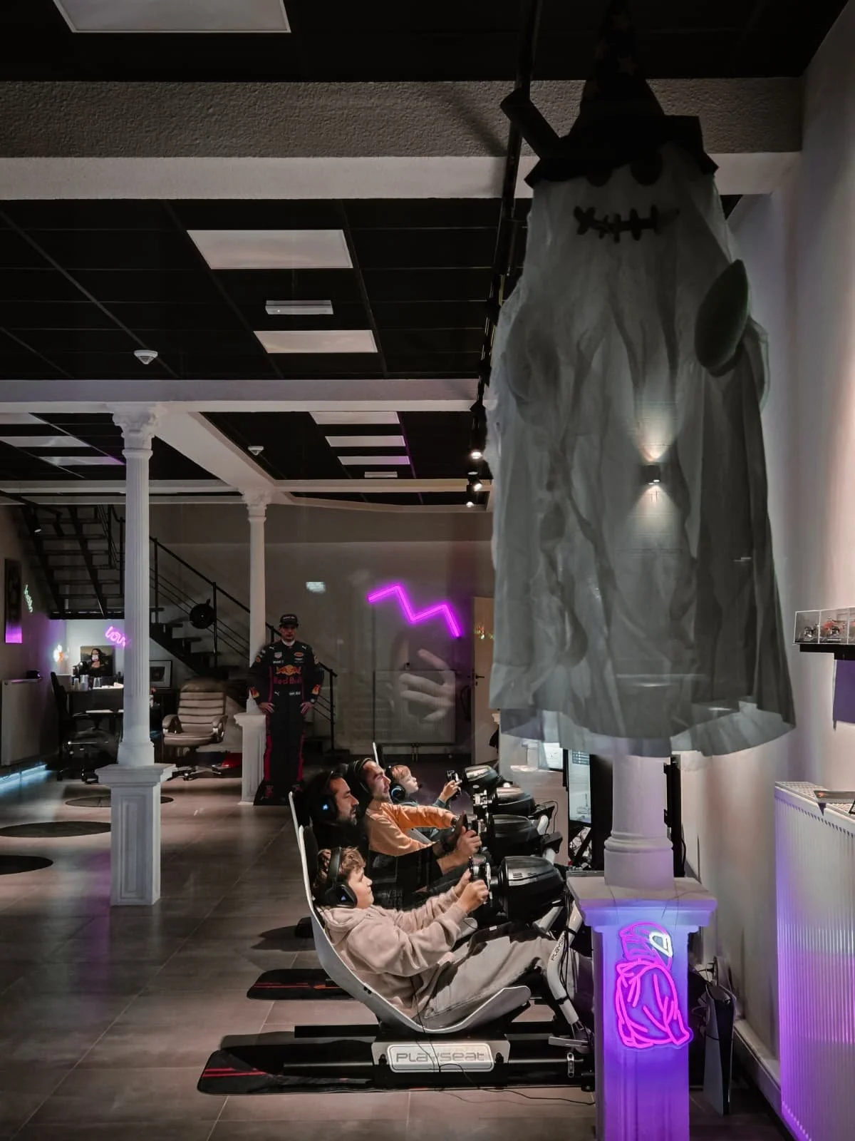 Quatre personnes jouent à un jeu de course sur des simulateurs de conduite dans un espace avec décor moderne et néons violets, avec une décoration d'Halloween suspendue au plafond.