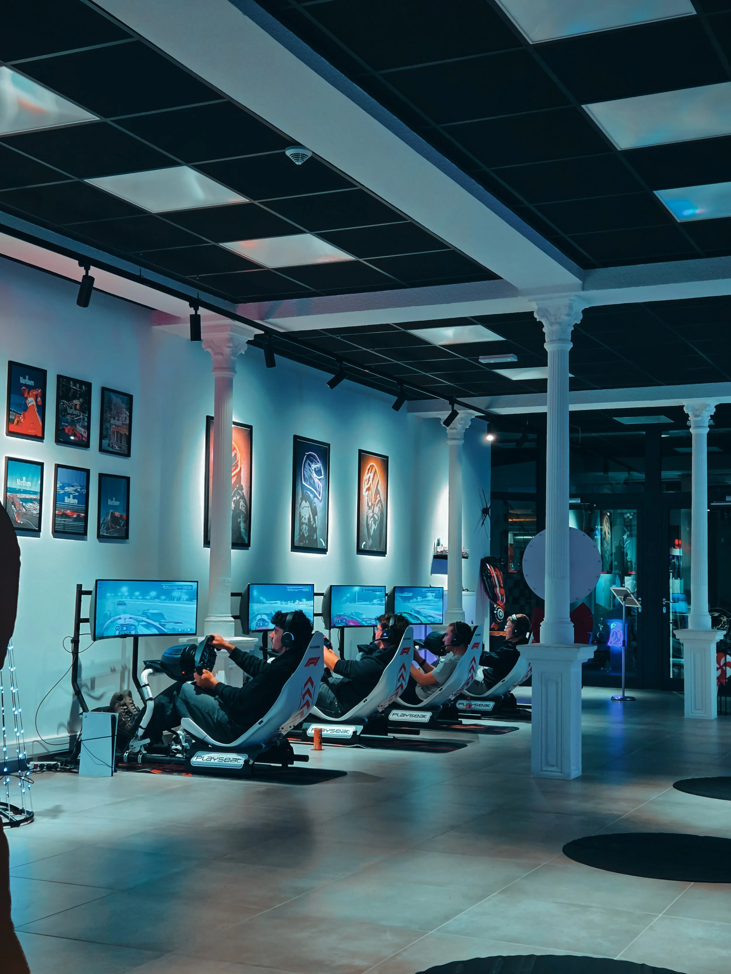 Salle d'arcade de course de voitures avec plusieurs personnes jouant en utilisant des volants et écrans, décoration moderne avec des posters sur le mur et colonnes blanches.