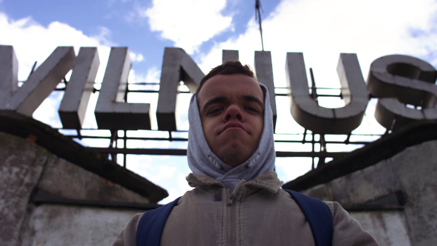 Young man with a hoodie and backpack standing in front of a sign that reads 'WILLITUS' against a cloudy sky.
