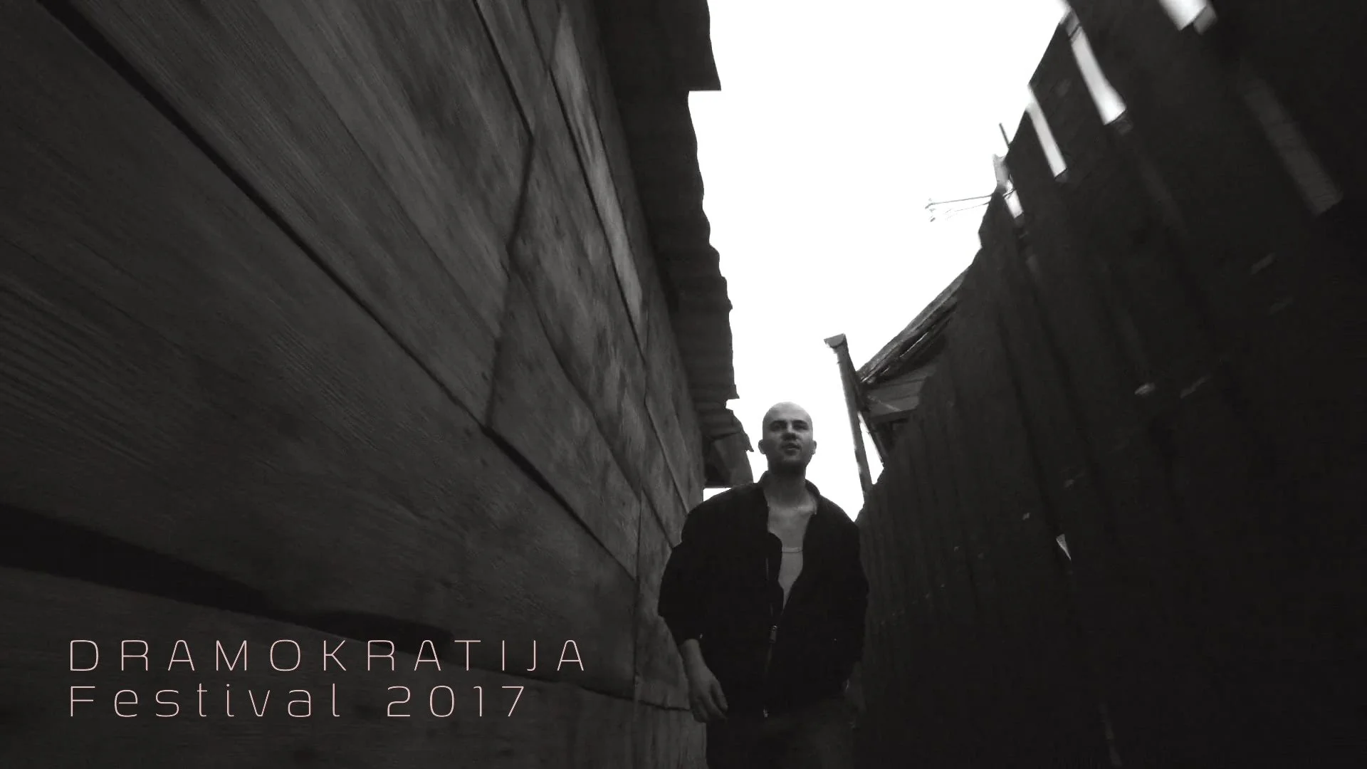 A black-and-white photo of a man walking between two narrow wooden walls or fences, with an overcast sky above. Text in the lower left corner reads 'DRAMOKRATIJA Festival 2017'.