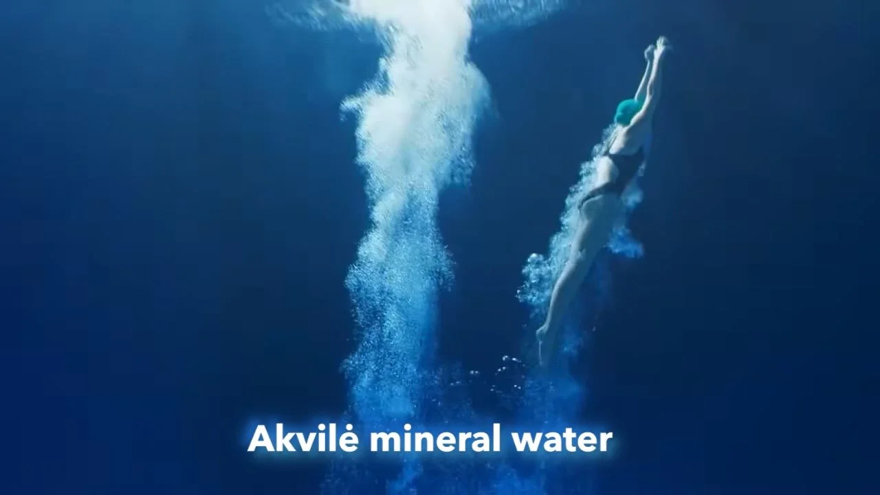 A woman swimming underwater in a pool with bubbles around her, wearing a swimsuit and swim cap, with the text 'Akvile mineral water' at the bottom.