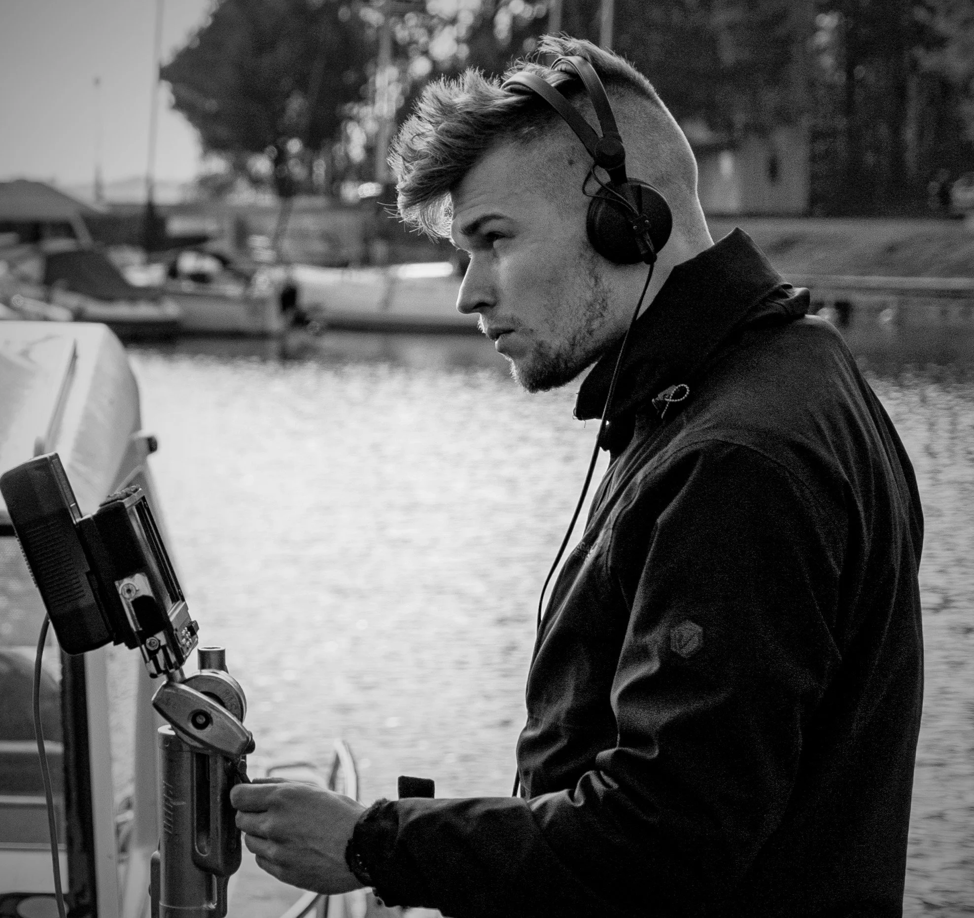 A young man with styled hair wearing headphones and a dark jacket, standing by a body of water with boats in the background, looking at equipment.