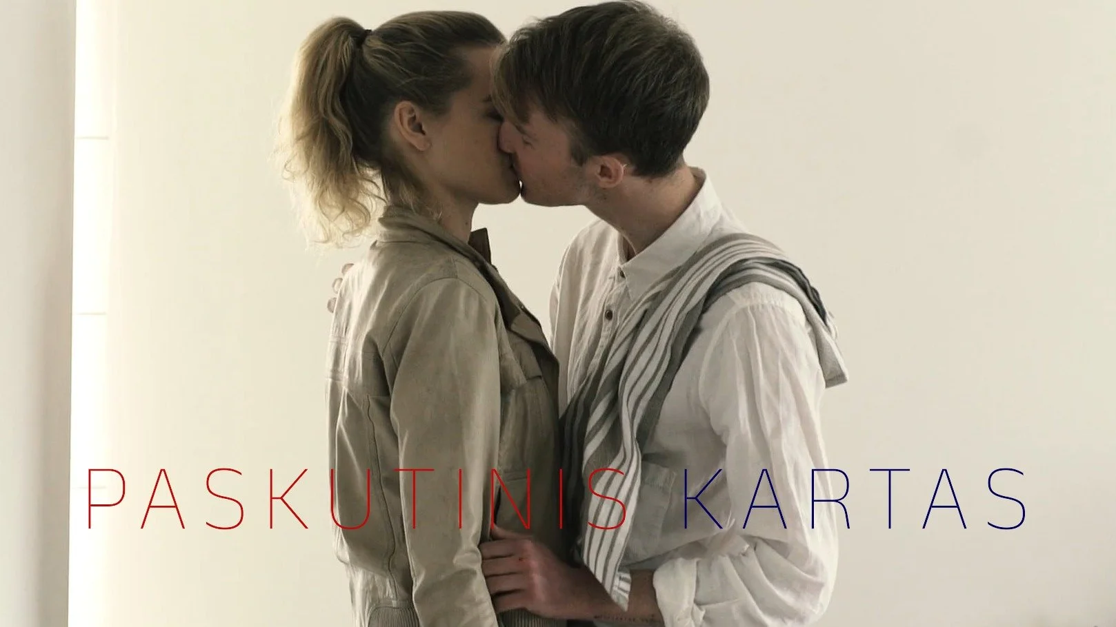 A man and woman are kissing in a minimalist setting with plain white walls. The woman has curly blonde hair tied back, and the man has short brown hair. The woman is wearing a beige jacket, and the man is wearing a white shirt with a striped sweater draped over his shoulders. The text "PASKUTINIS KARTAS" is overlaid in red and blue at the bottom of the image.