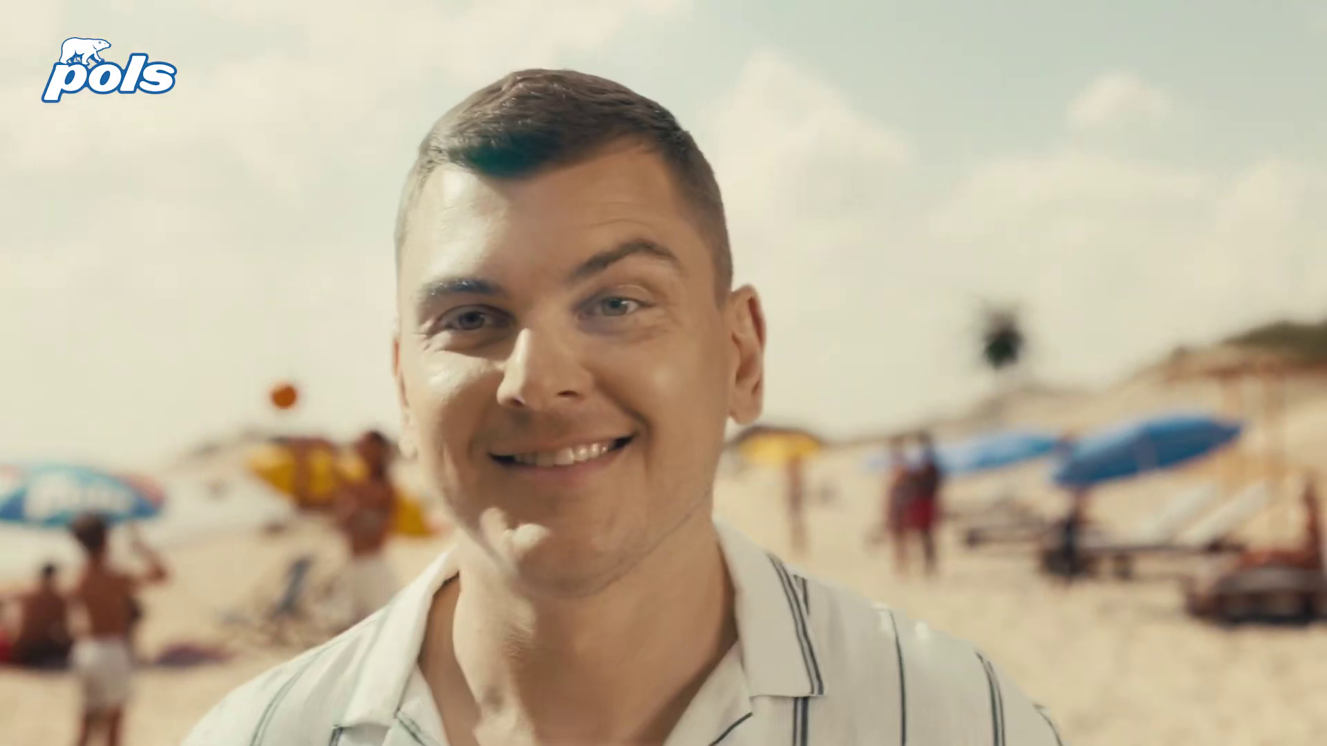 A young man smiling on a beach with colorful umbrellas and people in the background.