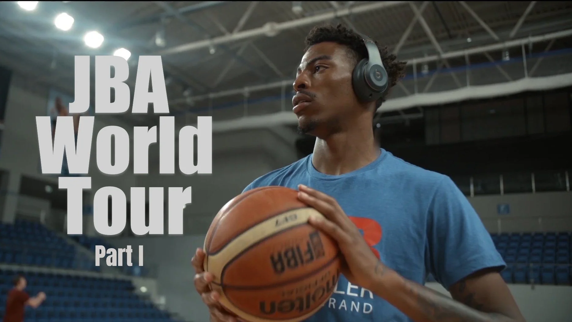 A young man with curly hair wearing a blue T-shirt and headphones holding a basketball in an indoor basketball court.