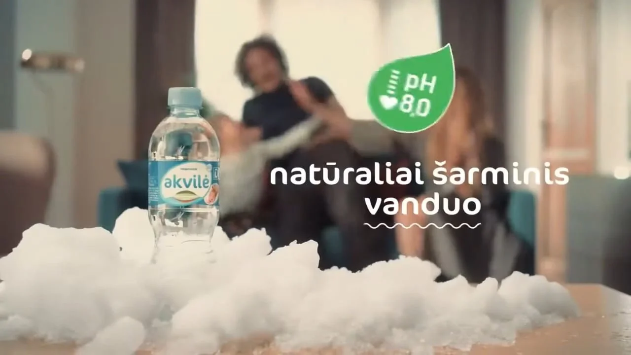 A bottle of Akvilė mineral water placed on ice with snow around it. The background shows a woman sitting on a couch.