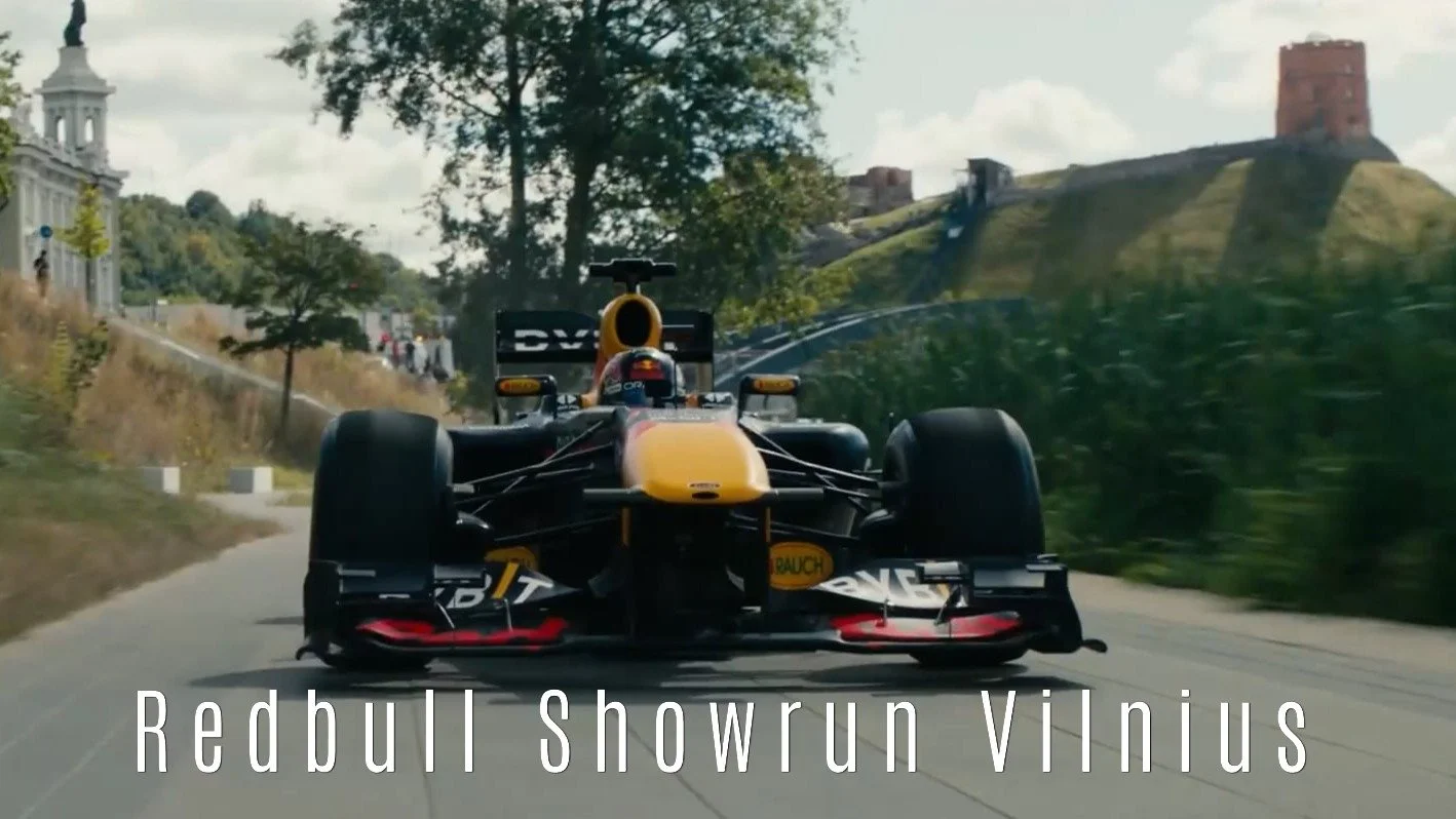 A Formula 1 race car driving on a narrow road in Vilnius, with the text 'Redbull Showrun Vilnius' overlaid.