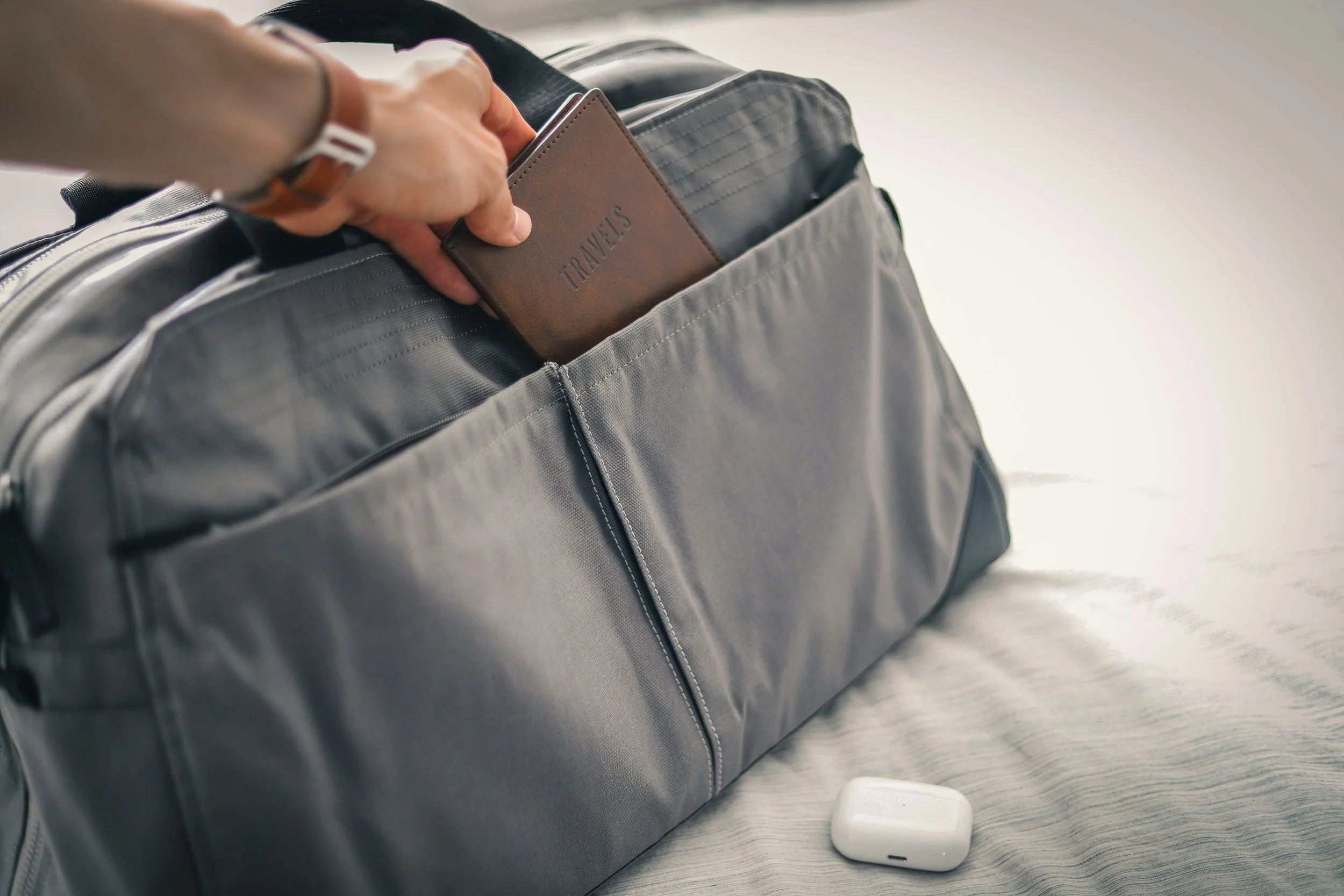 A gray travel bag on a bed with a person's hand placing a brown passport holder labeled "TRAVEL" into the outside pocket, and a white wireless earbud case on the bed.