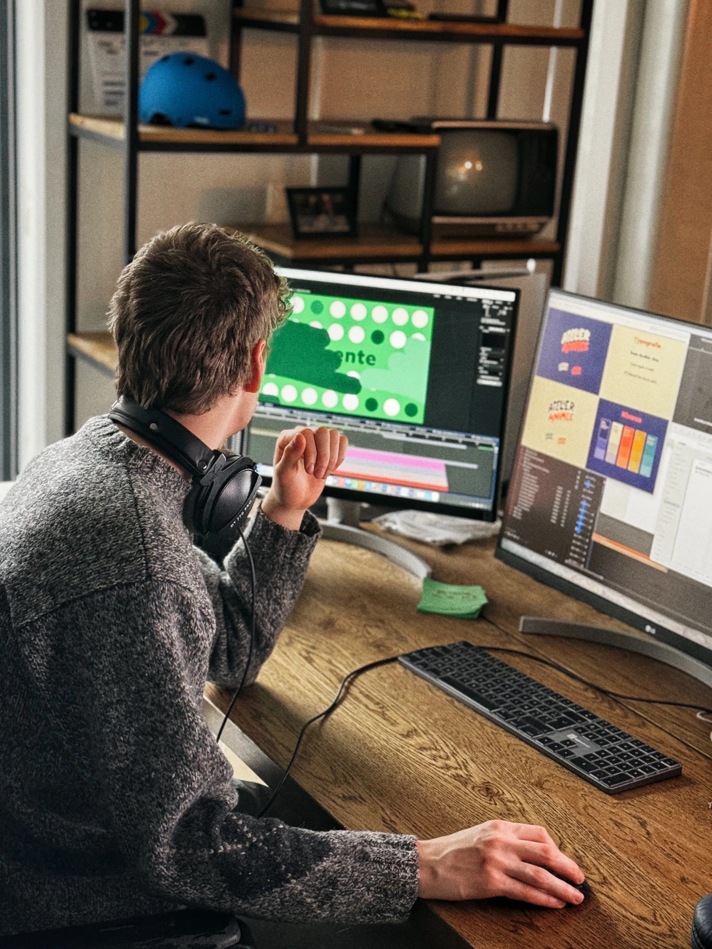Man werkt aan digitale grafische ontwerpen op twee beeldschermen, met een pc-toetsenbord en muis op een houten bureau, draagheadset hangt om zijn nek.
