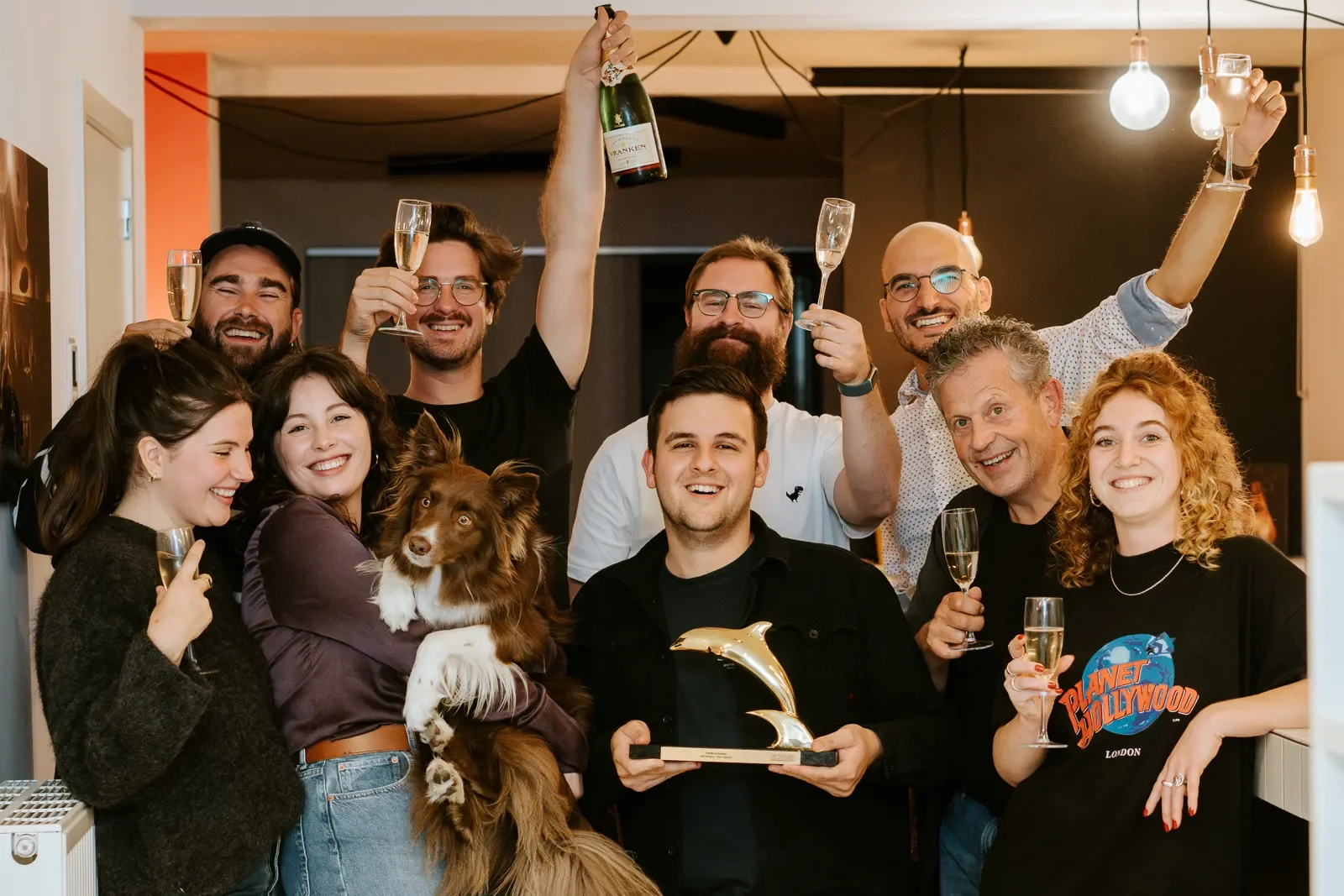 Een groep mensen feestend met champagne, één persoon houdt een award vast, er is een hond en ze lachen allemaal.