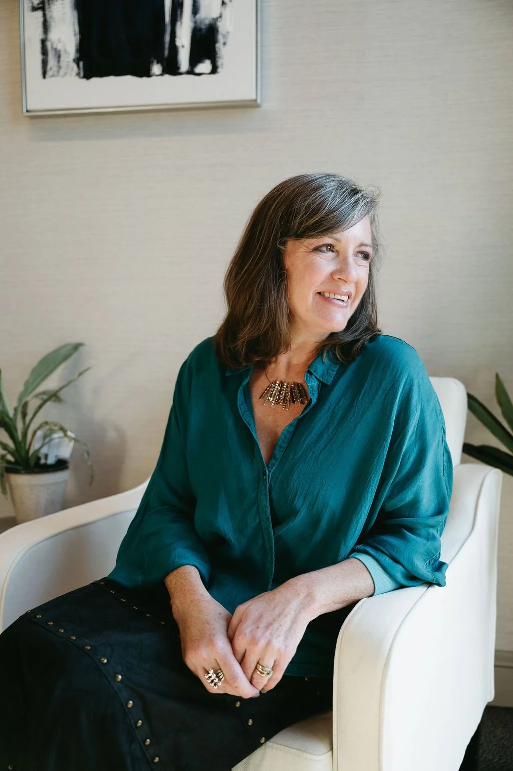 A woman sitting in a cream-colored armchair, smiling and looking to her right. She has shoulder-length brown hair and is wearing a teal blouse, a statement necklace, and rings on her fingers. There are potted plants and framed artwork on the wall behind her.