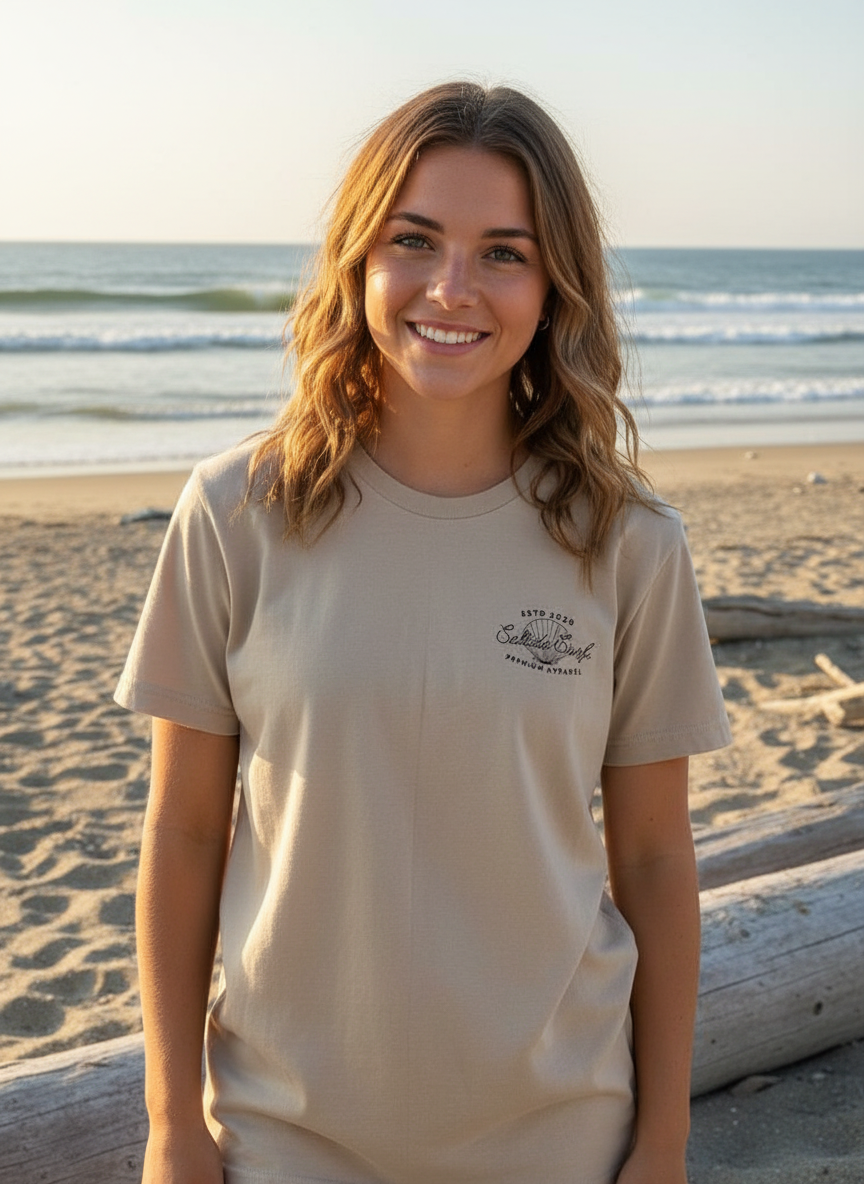 A young woman with wavy, shoulder-length brown hair and fair skin smiling at the camera on a beach, with the ocean and gentle waves in the background during sunset.