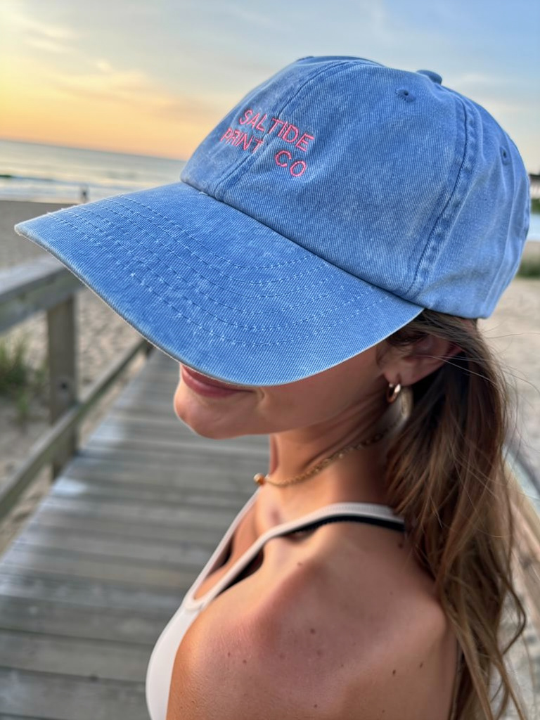 A woman wearing a blue baseball cap with pink embroidery reading 'Saltide Print Co' stands on a wooden pier at the beach during sunset.