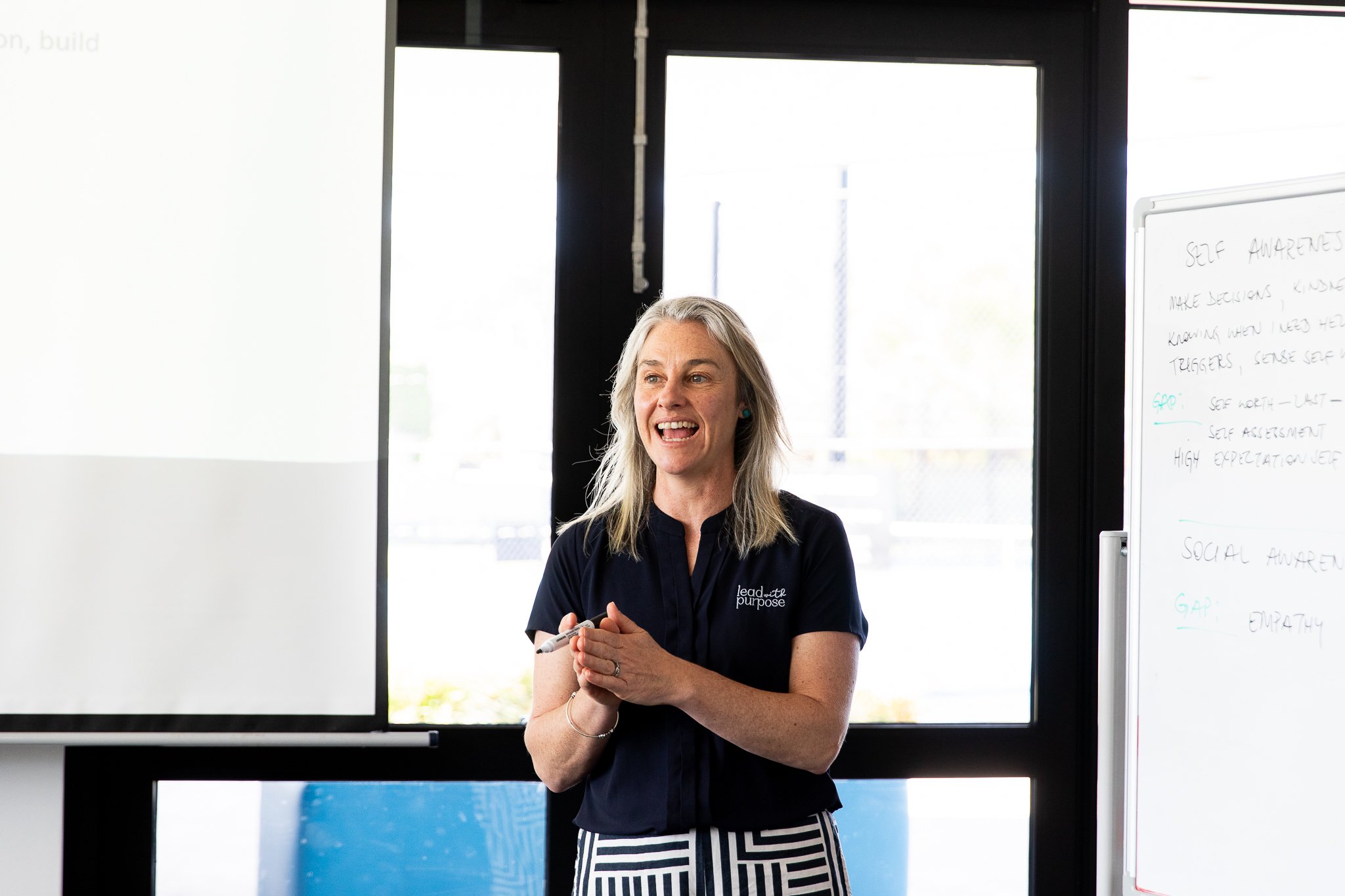Karissa Cameron, a woman, wearing a navy shirt with a logo, talking in front of a whiteboard and windows.